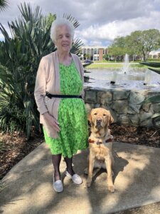 Karen stands in a landscaped area near a fountain with yellow Lab guide dog Gadget sitting beside her