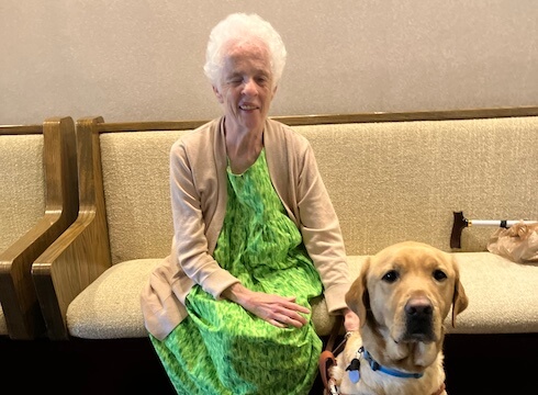 Karen sits on a padded bench indoors with her hand on yellow Lab guide dog Gadget's back