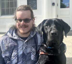 Justin sits outside on a sunny day with black guide dog Tootsie for their team portrait