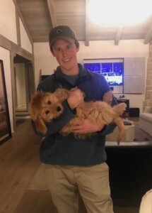 Alex stands inside a living area holding a small yellow golden retriever-type dog in his arms smiling at camera