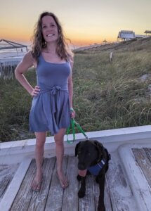Ashley wears a lavender sundress standing on a wooden staircase with black lab guide dog Lolly in a beachy  area at sunset