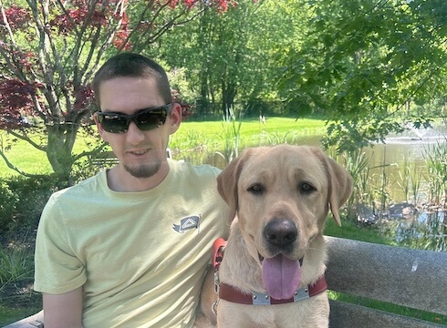 Chris and yellow lab guide dog Nigel sit close on an outdoor bench against backdrop of greenery and the pond
