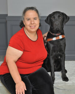 Judy is in a red shirt and black pants sitting next to black Lab guide dog Lyons in their portrait
