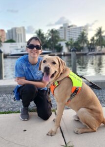 Mariana is on one knee next to yellow lab guide Einstein in an urban setting in front of a body of water