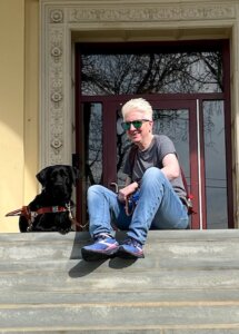 Pam sits with black Lab guide dog Mary at the top of a long flight of cement steps leading to an ornate entrance