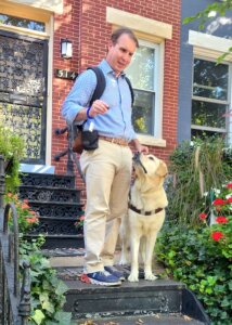 Pat Leahy stands with yellow Lab guide dog Hogan on steps with flowers growing on either side leading to a brick residence