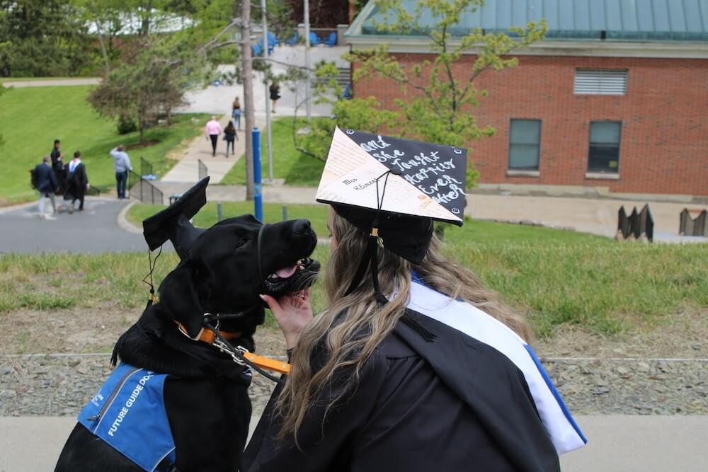 Pup Imus and his raiser Emily seen from behind both wearing graduation caps on a hill overlooking Hartwick campus