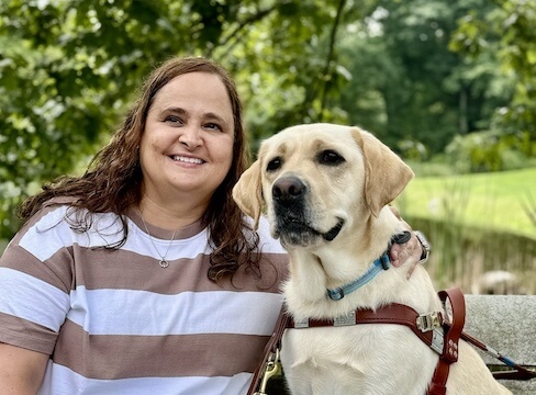 Aimee and yellow guide dog Bryna sit on a stone bench against a background of greenery