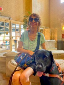 Carol and black Lab guide dog Ezra sit in a lobby with glass doors and stone benches for their team portrait