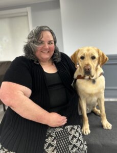 Heather and yellow Lab guide dog Darby sit together indoors for their team portrait