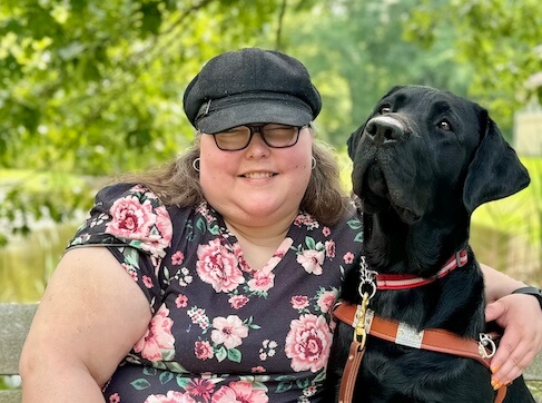 Andrea leans in to embrace black lab guide dog Ingalls while sitting outdoors on a bench for their team portrait
