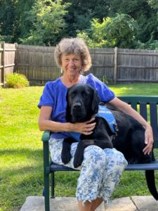 Maggee sits on an outdoor bench smiling as black lab guide dog Wegman lies across her lap with head up.