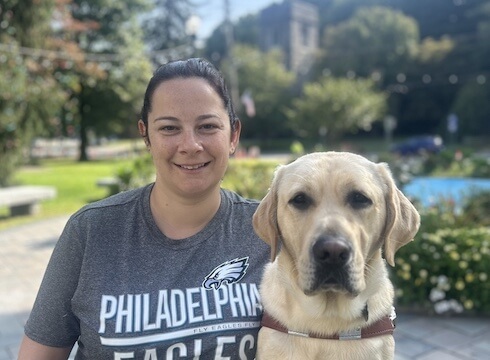 Michelle and yellow guide dog Albert sit on a stone bench in a park like setting for their team portrait