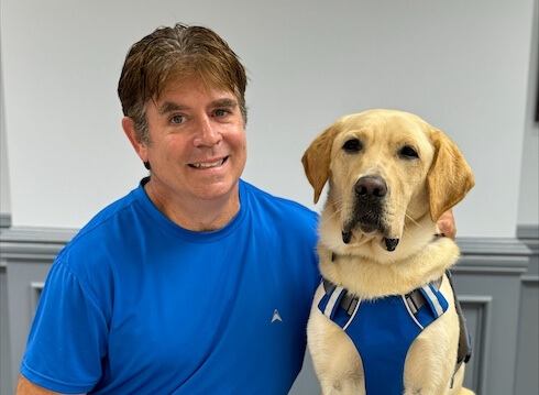 Rob and yellow lab guide dog Maguire sit side by side for their indoor team portrait.