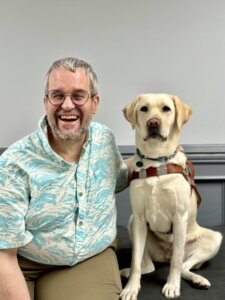 Richard and yellow Lab guide dog Baja sit side by side for their indoor team portrait