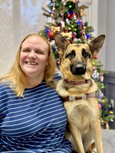 Dawn and German Shepherd guide dog Gussie sit for team portrait with festive holiday tree behind their heads.