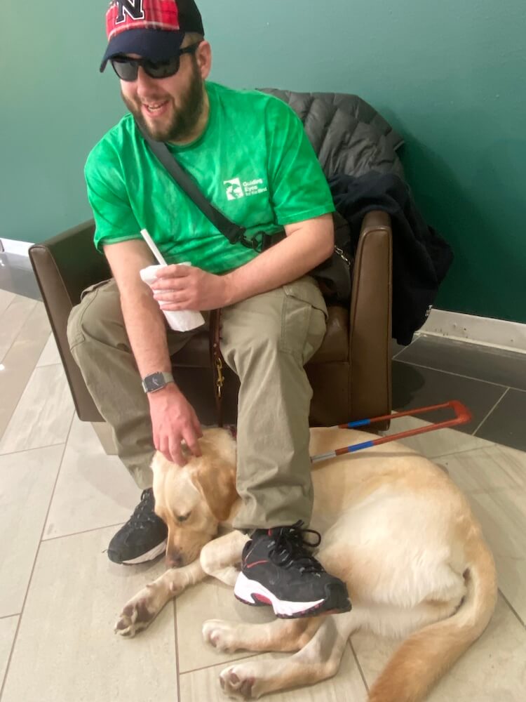 Dustin sits in a cushioned chair and reaches down to pet guide Queen lying at his feet