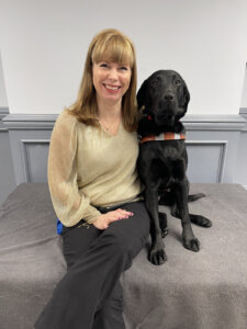 Noella sits close to black Lab guide dog Doll indoors for their team portrait