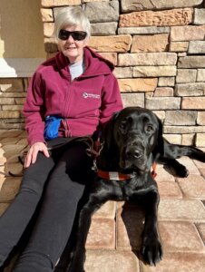 Susan sits on a tiled area with her back against a brick wall as her black Lab guide dog Cara is in a down next her outstretched legs.