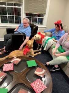Two yellow labs wearing red accessories touch noses while their handlers pet their backs.