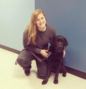 Shannon is on one knee with her arm around a black lab. 