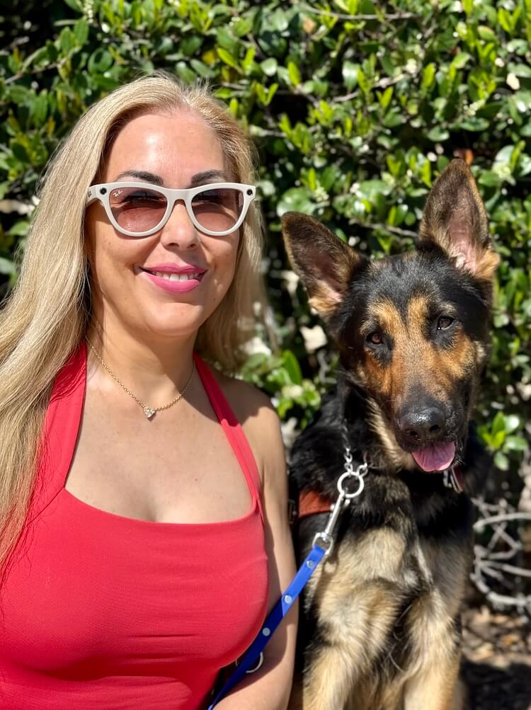 Irma looks towards camera and sits with German Shepherd guide dog Wonder who head tilts slightly in a bright sunny location