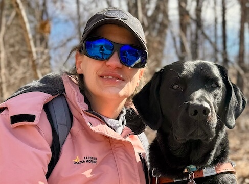 Jessica in a pink jacket sits close to black Lab guide dog Jess in front of late winter trees without leaves