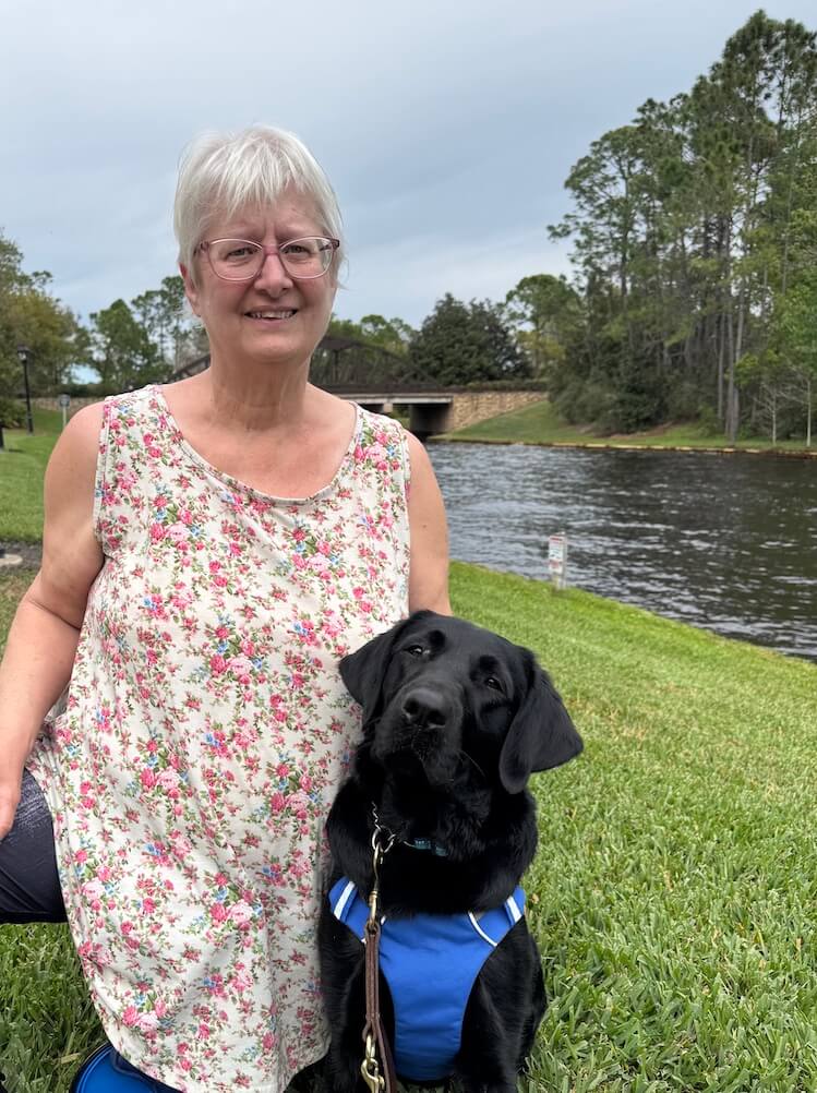 Mary kneels in a brightly lit area next to a river flowing under a bridge with black Lab guide dog Indra sitting next to her