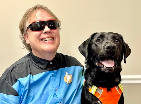 Todd and black Lab guide dog Edsel sit side by side with big smiles for their indoor team portrait