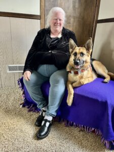 Cindy sits on bench draped in dark purple next to her German Shepherd guide dog Gretel who lies beside her with a proud happy expression