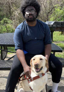 Anthony sits at a black metal outdoor table with his yellow guide dog Clancey sitting at his feet