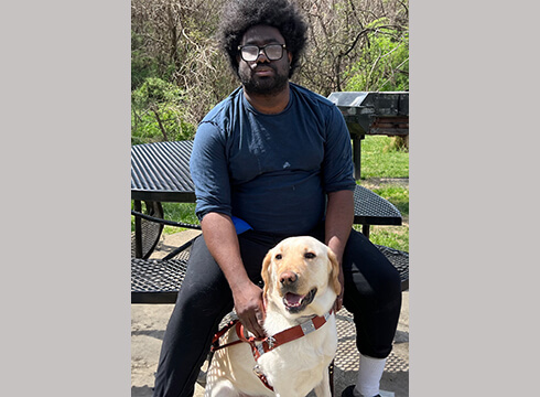 Anthony sits at a black metal outdoor table with his yellow guide dog Clancey sitting at his feet