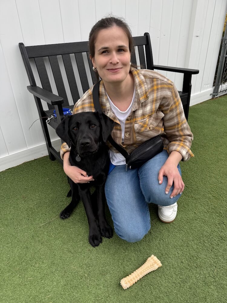 Maddie kneels down gently holding black Lab Jackie beside her on the turf of the dog runs