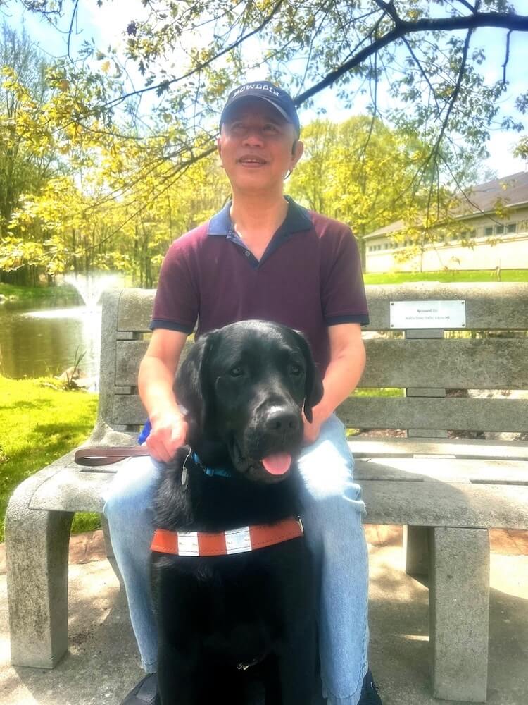 Allen and black guide dog Rex are on a park bench on the Yorktown campus for their team portrait