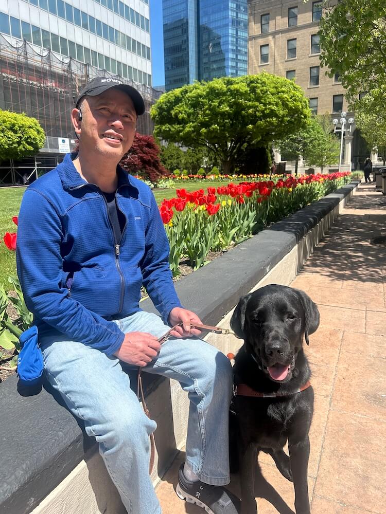 Allen sits on a retaining wall with a long line of red tulips beyond him and guide Rex