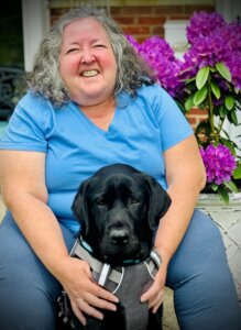 Ann with black guide dog Iowa sitting directly in front of her by a pot of purple flowers in front of a building for their outdoor team portrait