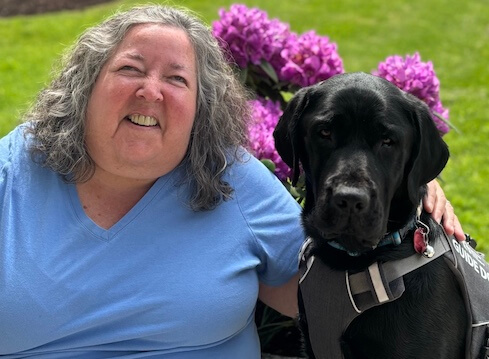 Ann and black guide dog Iowa sit side by side near a purple flowering bush