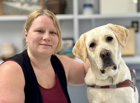 Brittany and yellow guide dog Pearse whose head tilts towards hers, sit indoors for their team portrait.