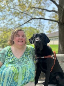 Kate and black guide dog Eagle are on a park bench on the Yorktown campus for their team portrait