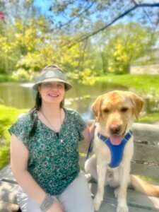 Leslie and yellow guide dog Othello are on a park bench with the pond and fountain of Yorktown campus behind them in their team portrait