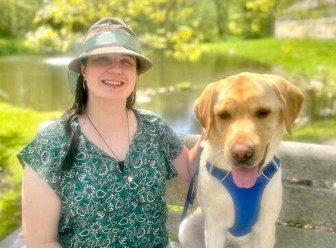 Leslie and yellow guide dog Othello are on a park bench with the pond and fountain of Yorktown campus behind them in their team portrait