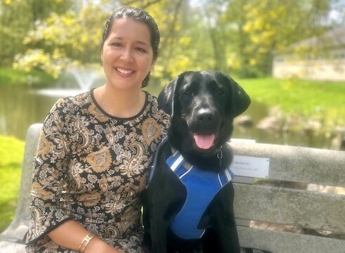 Rebekah and black guide dog Kokomo are on a park bench on the Yorktown campus for their team portrait