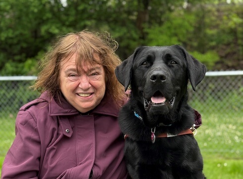 Rose and black Lab guide dog Vintage sit side by side on an outdoor wood bench - a happy expression on both faces