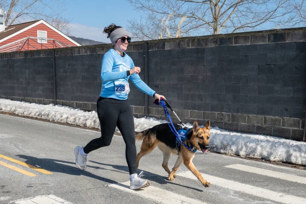 Woman runs outdoors with a German Shepherd guide dog in blue Unifly harness.