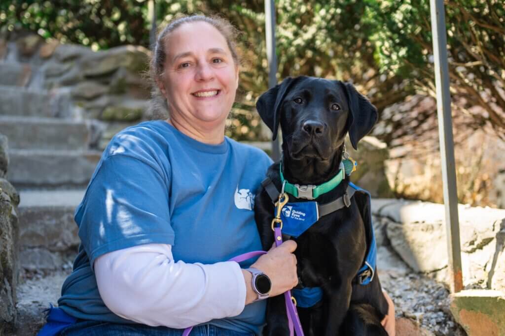 Woman in blue shirt smiles while sitting outdoors with a Black Lab in blue Future Guide Dog vest.