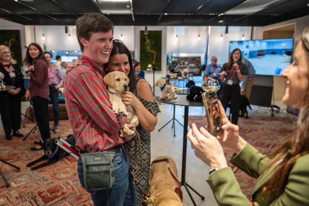 Smiling couple holds a Yellow Lab puppy as another guest takes their photo at an event.