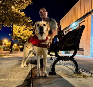 Guide Lindsey stands in front of the bench handler Anthony is sitting on, within a paved area lit by streetlights and lined by young trees