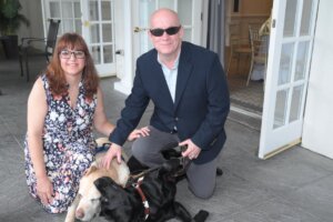 Graduate Bill Smith and his wife kneel, while smiling for the camera. Guide dog Quidditch and retired guide Ed lay on the ground between them. 