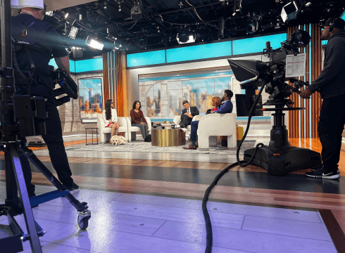 A wide shot of the CBS Mornings studio set, showing the full backdrop and several cameras as the interview is about to begin. On the left side of the set is Marin and Rebekah, each with their respective guide dog. On the right side are the three anchors, Gayle, Tony, and Nate, each with a black lab puppy on their lap.