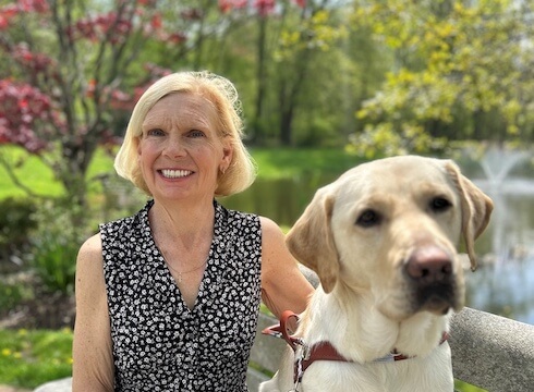 Deborah and yellow guide dog Keeper are on a park bench with the pond and fountain of Yorktown campus behind them in their team portrait